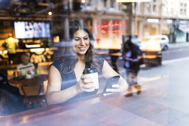 Happy young woman sitting with coffee cup and using mobile phone in cafe - WPEF08604