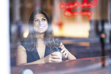 Happy woman sitting with coffee cup and seen through glass - WPEF08605