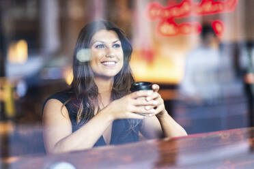 Smiling young woman sitting with coffee cup and seen through glass - WPEF08612