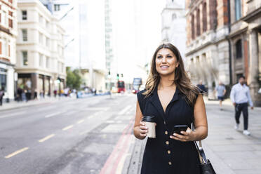 Lächelnde Frau mit Kaffeetasse und Smartphone auf einem Fußweg in der Stadt - WPEF08622
