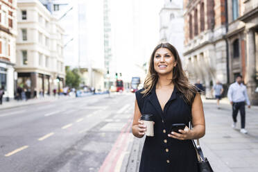 Smiling woman standing with coffee cup and smart phone on footpath in city - WPEF08622