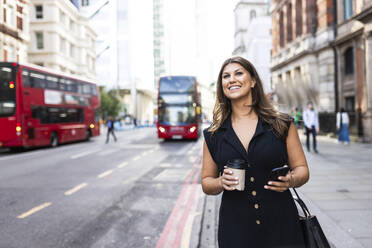 Lächelnde junge Frau mit Kaffeetasse und Smartphone auf einem Fußweg in der Londoner Innenstadt - WPEF08624
