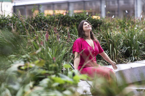 Smiling young woman sitting with eyes closed near plants - WPEF08652