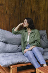Nachdenkliche Frau sitzt auf dem Sofa vor einer Holzwand zu Hause - SEAF02257
