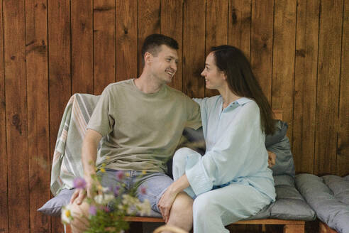 Romantic couple looking at each other sitting on sofa in front of wooden wall at home - SEAF02262