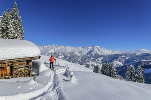 Ältere Frau beim Skifahren vor einer verschneiten Hütte an einem sonnigen Tag, Schwalbenwand, Dientener Berge, Salzburg, Österreich - ANSF00827