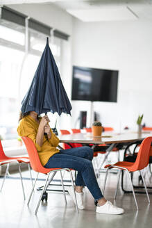 Geschäftsfrau mit gefaltetem Regenschirm, der das Gesicht in einem kreativen Büro bedeckt - KNSF10041