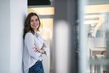 Smiling confident businesswoman with arms crossed leaning on wall in office - KNSF10056