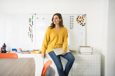 Thoughtful businesswoman with laptop sitting on cabinet in office - KNSF10057