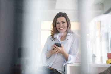 Smiling businesswoman with smart phone sitting at desk in creative office - KNSF10059