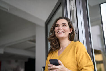 Nachdenkliche Geschäftsfrau mit Smartphone am Fenster im Büro - KNSF10066