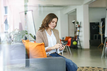 Businesswoman using smart phone sitting on sofa in office - KNSF10073