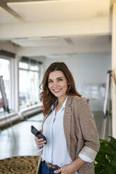 Lächelnde Geschäftsfrau mit Mobiltelefon im Büro stehend - KNSF10079