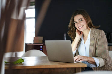 Lächelnde Geschäftsfrau mit Laptop am Schreibtisch im Büro - KNSF10085