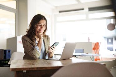 Smiling businesswoman using mobile phone at laptop desk in office - KNSF10088