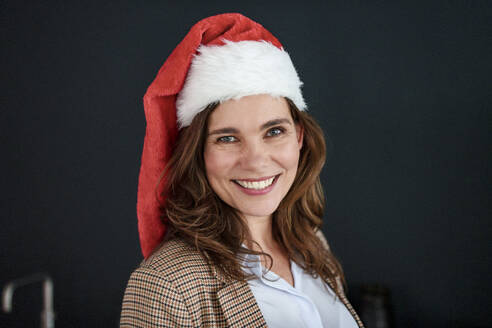 Cheerful businesswoman in santa hat in front of black background - KNSF10098