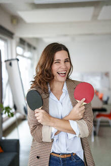 Fröhliche Geschäftsfrau hält Tischtennisschläger in einem kreativen Büro - KNSF10103