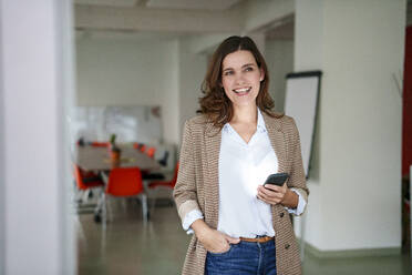 Lächelnde reife Geschäftsfrau mit Handy in einem kreativen Büro - KNSF10119