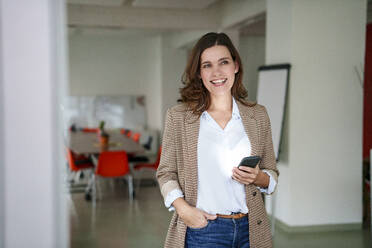 Smiling mature businesswoman holding mobile phone in creative office - KNSF10119