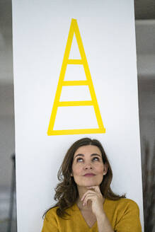 Nachdenkliche kreative Geschäftsfrau, die ein Pyramidenschild an der Wand betrachtet - KNSF10127