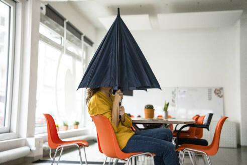 Kreative Geschäftsfrau mit gefaltetem Regenschirm, der das Gesicht am Arbeitsplatz bedeckt - KNSF10135