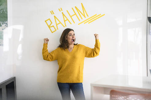 Kreative Geschäftsfrau, die ihre Muskeln vor einer weißen Wand mit Text im Büro anspannt - KNSF10157