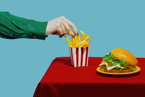 Hand einer Frau, die Pommes frites vom Tisch aufhebt, vor blauem Hintergrund - RDTF00062