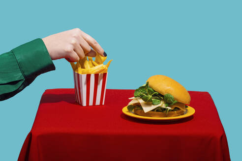 Frau greift nach Pommes frites in der Nähe von Cheeseburger auf Tisch vor blauem Hintergrund - RDTF00069