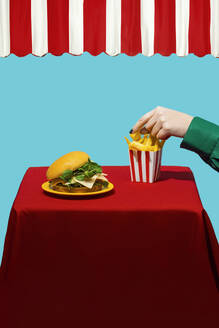 Hand of woman reaching for french fries near burger on table against blue background - RDTF00070