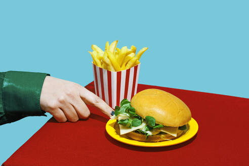 Hand of woman touching plate of cheeseburger on table against blue background - RDTF00071