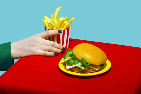 Woman picking up french fries near burger on table against blue background - RDTF00072