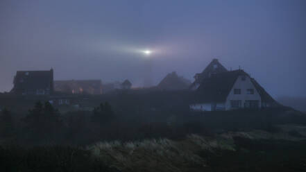 Deutschland, Schleswig-Holstein, Hornum, Dorfhäuser in der nebligen Morgendämmerung mit leuchtendem Leuchtturm im Hintergrund - KEBF02858