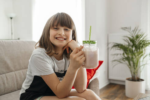 Happy girl holding mason jar and sitting on sofa at home - ELMF00112