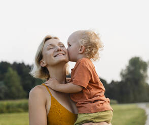 Boy kissing happy mother in meadow - NDEF01616