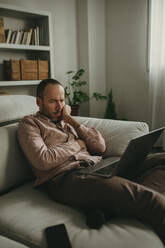 Thoughtful freelancer sitting on sofa and looking at laptop in home office - DMGF01283