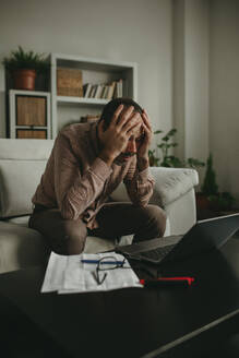 Tired freelancer sitting with head in hands near laptop at home office - DMGF01294