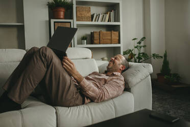 Cheerful man lying down and watching movie on laptop at home - DMGF01297