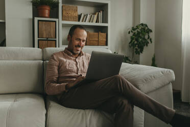 Smiling mature man watching movie on laptop at home - DMGF01298