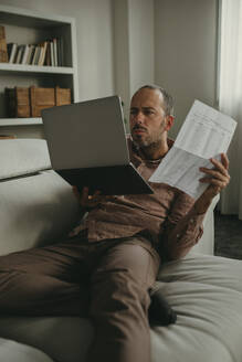 Serious freelancer holding document and looking at laptop in home office - DMGF01309