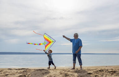 Senior man and grandson flying kite near sea at beach - MBLF00295