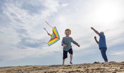 Playful boy with grandfather flying kite at beach - MBLF00298