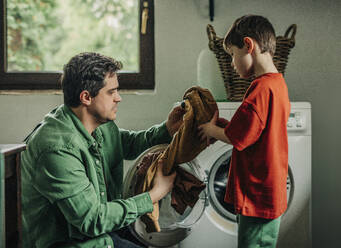 Father and son removing clothes from washing machine at home - VSNF01751