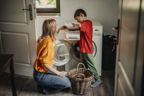 Son pouring detergent in washing machine with mother crouching at home - VSNF01761