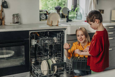 Mutter und Sohn halten schmutzige Löffel in der Nähe der Spülmaschine in der Küche zu Hause - VSNF01768