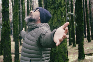 Carefree man with arms outstretched leaning on tree trunk in forest - SIPF02903