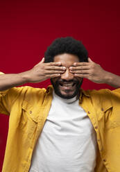 Smiling man covering eyes with hands against red background - VRAF00466