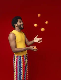 Glücklicher Mann jongliert mit Orangen vor rotem Hintergrund - VRAF00473