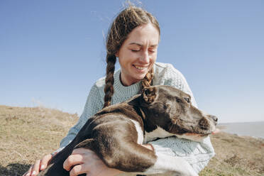 Lächelnde Frau verbringt Freizeit mit Hund auf einem Hügel an einem sonnigen Tag - KVBF00057