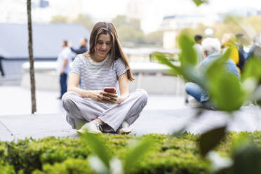 Lächelnde junge Frau, die ein Smartphone benutzt und in der Nähe von Pflanzen sitzt - WPEF08659