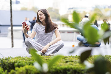 Junge Frau, die ein Smartphone benutzt und in der Nähe von Pflanzen sitzt - WPEF08660